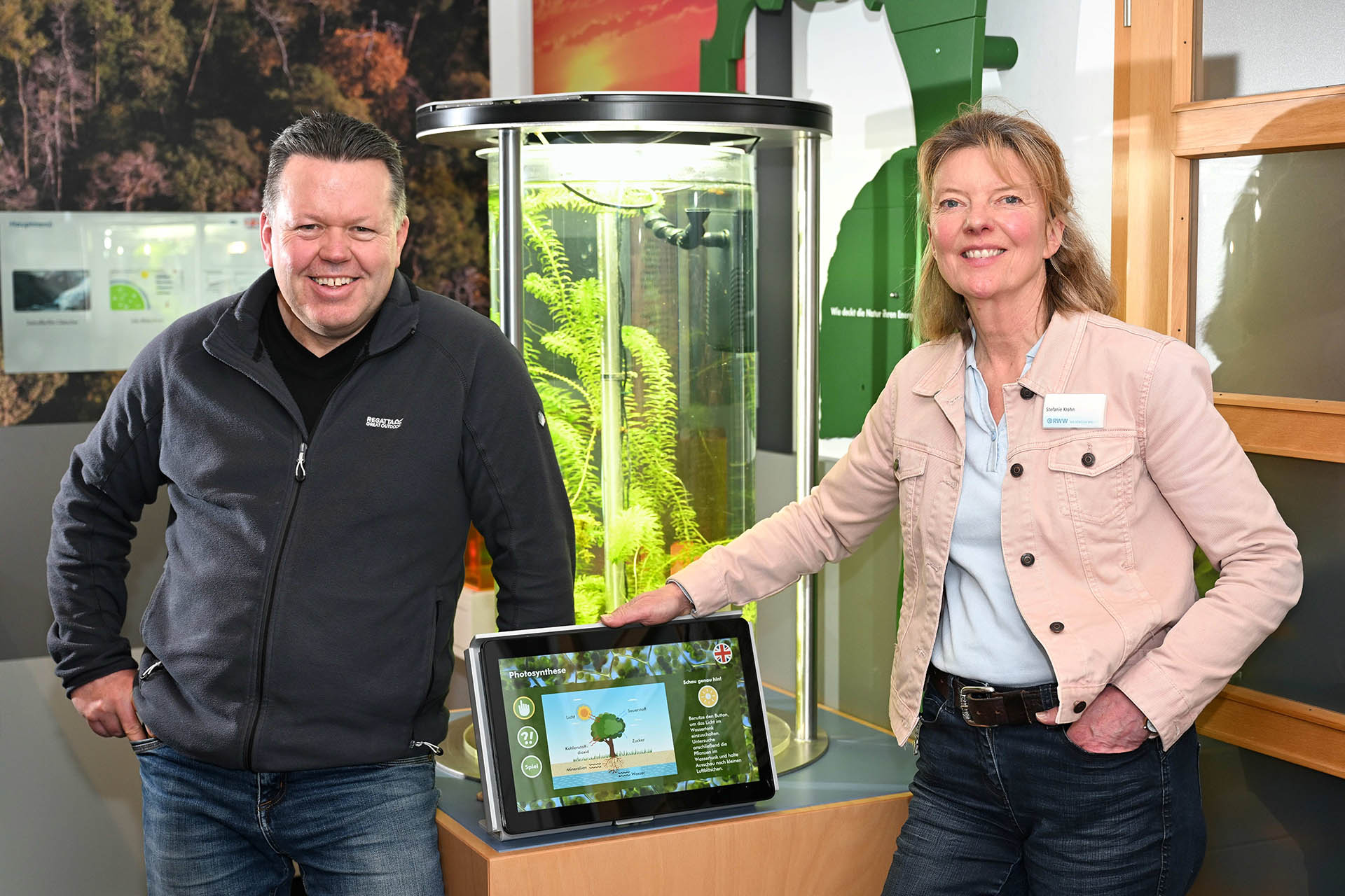 Ein Mann und eine Frau halten ein Laptop. Im Hintergrund sieht man einen Teil einer Naturausstellung.